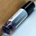 A small cylindrical perfume or roll-on bottle labeled 'UPSCENTS JIMMY CHILL' resting diagonally on a light surface next to a wooden background.